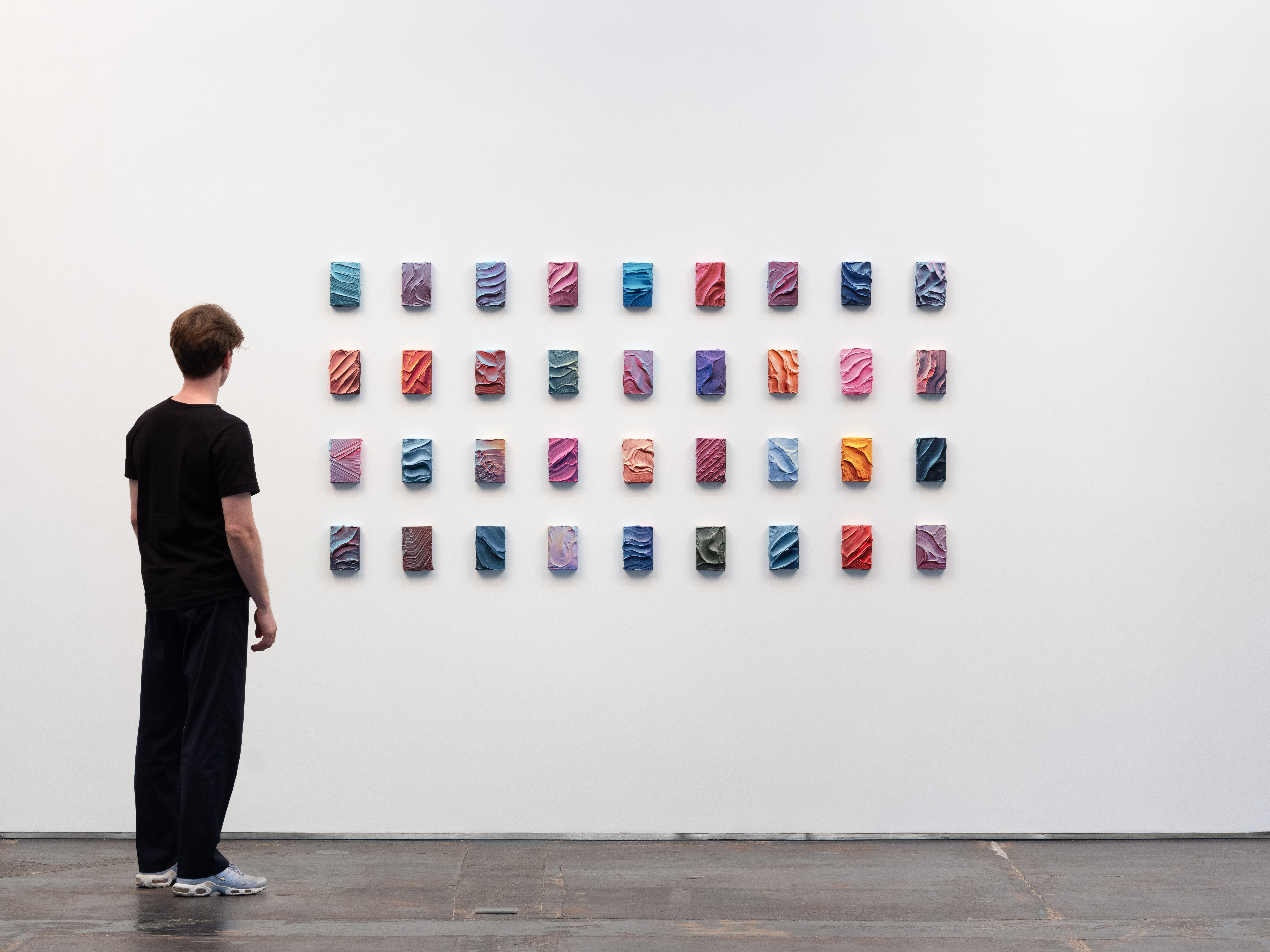 Ruben Benjamin, After The Dust Settles, 2026 — installation view at Gallery Sonder showing a grid of small, colorful mixed media works arranged on a white wall, with a viewer standing nearby for scale, emphasizing variation, repetition, and material detail. Gallery Sonder, Orange County.