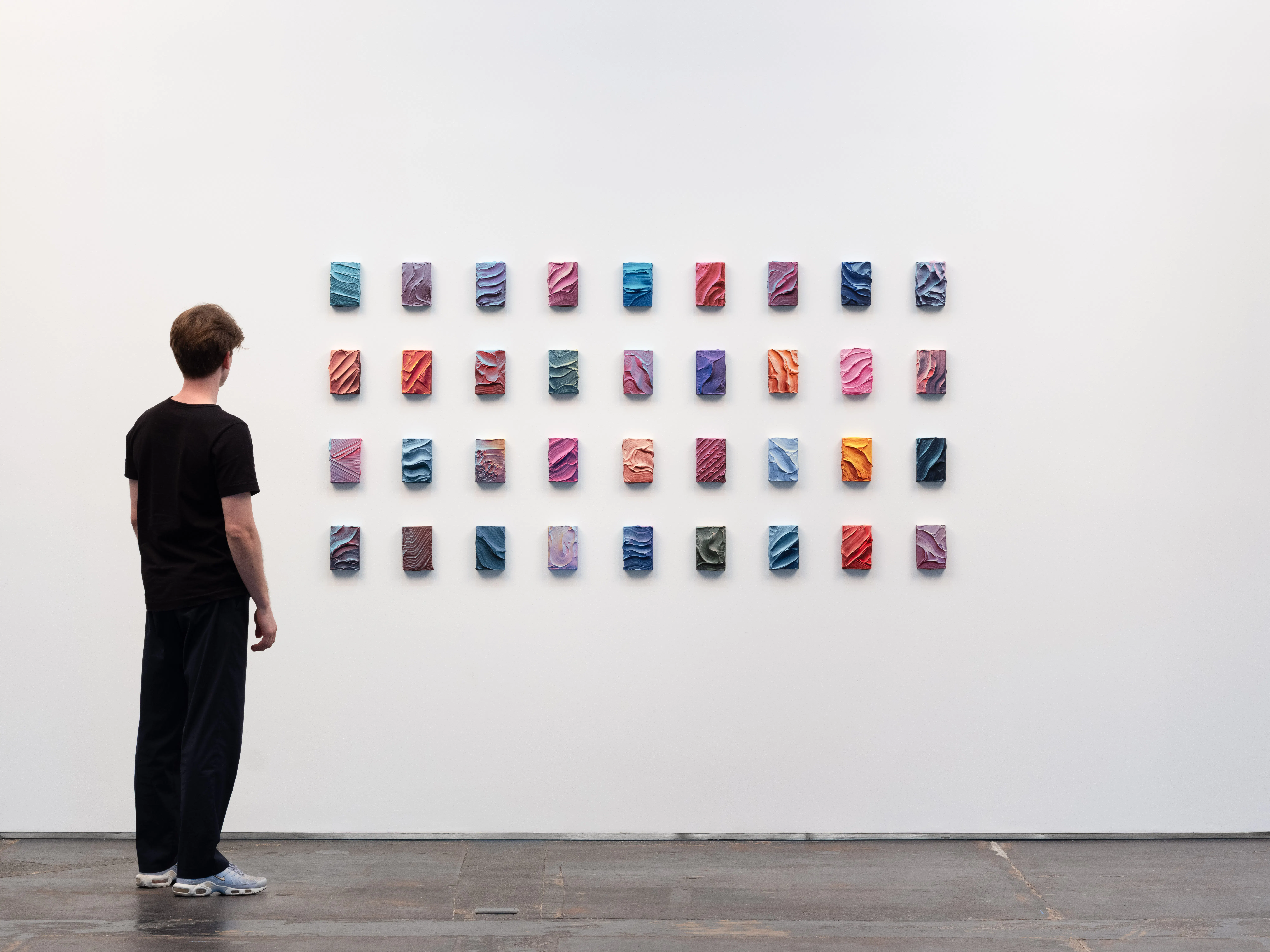 Ruben Benjamin, After The Dust Settles, 2026 — installation view at Gallery Sonder showing a grid of small, colorful mixed media works arranged on a white wall, with a viewer standing nearby for scale, emphasizing variation, repetition, and material detail. Gallery Sonder, Orange County.