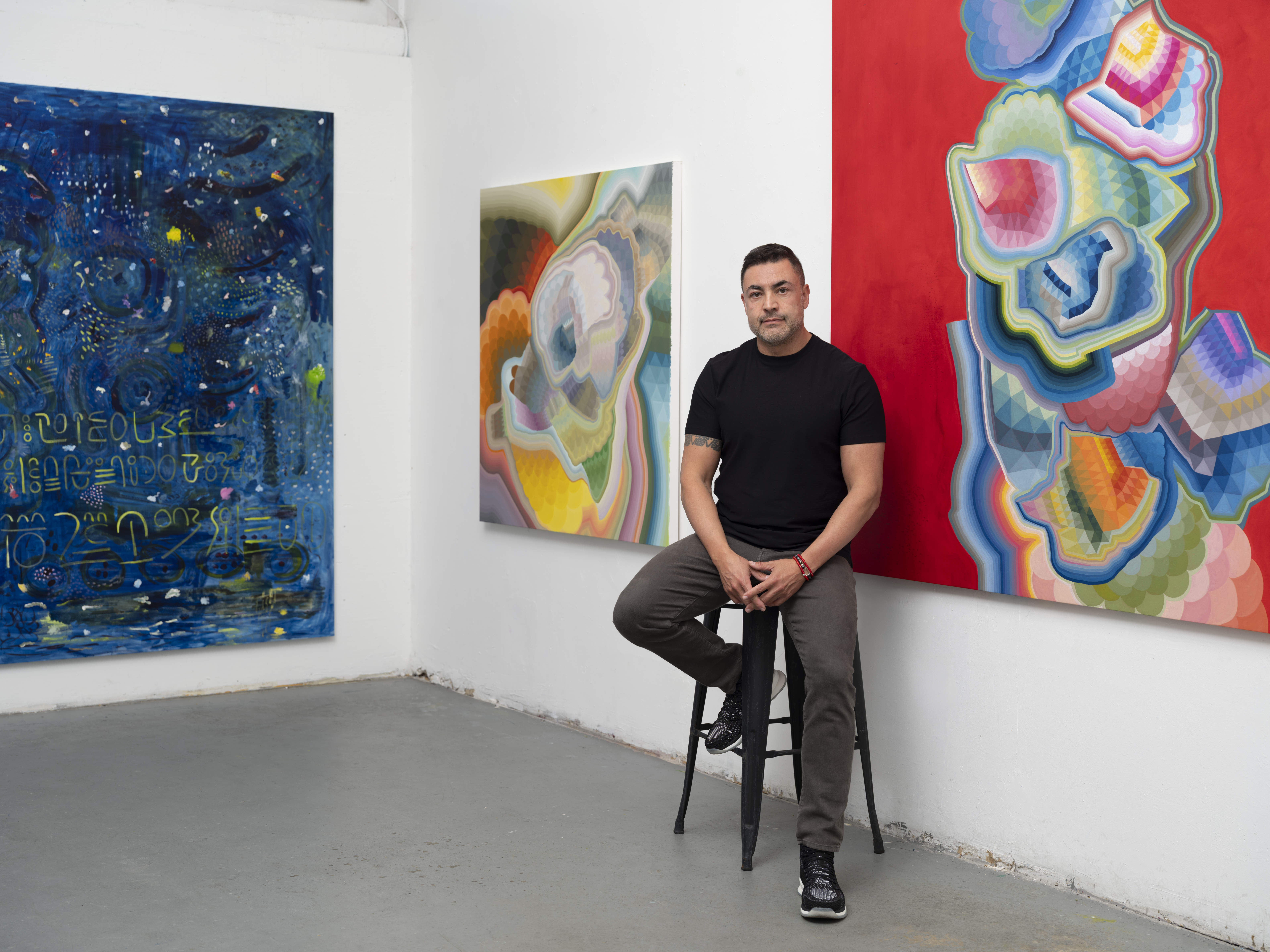Moncho in his studio, seated on a stool between large-scale abstract paintings, with colorful geometric compositions visible on surrounding walls, emphasizing the artist within his working environment.