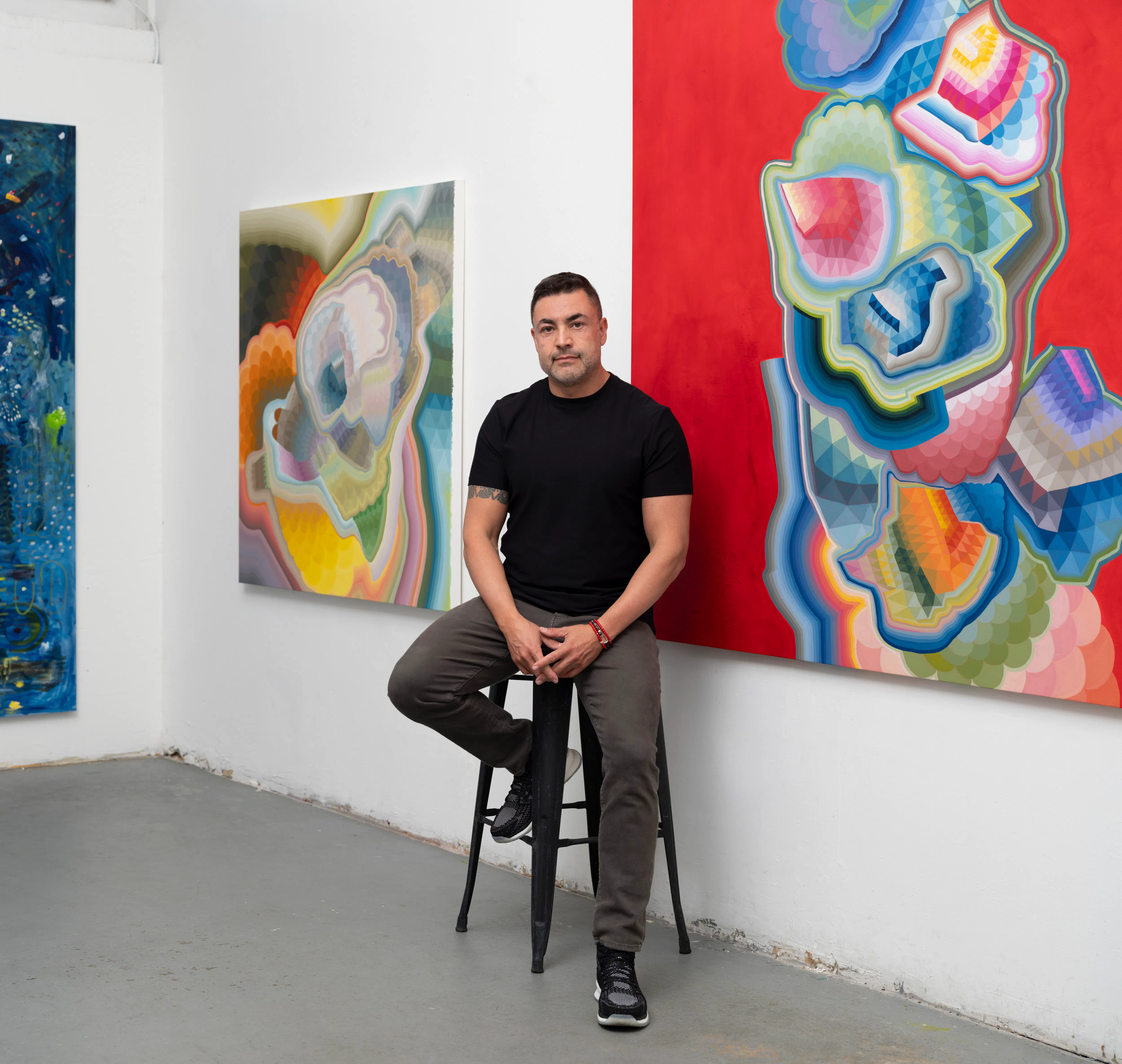Moncho in his studio, seated on a stool between large-scale abstract paintings, with colorful geometric compositions visible on surrounding walls, emphasizing the artist within his working environment.