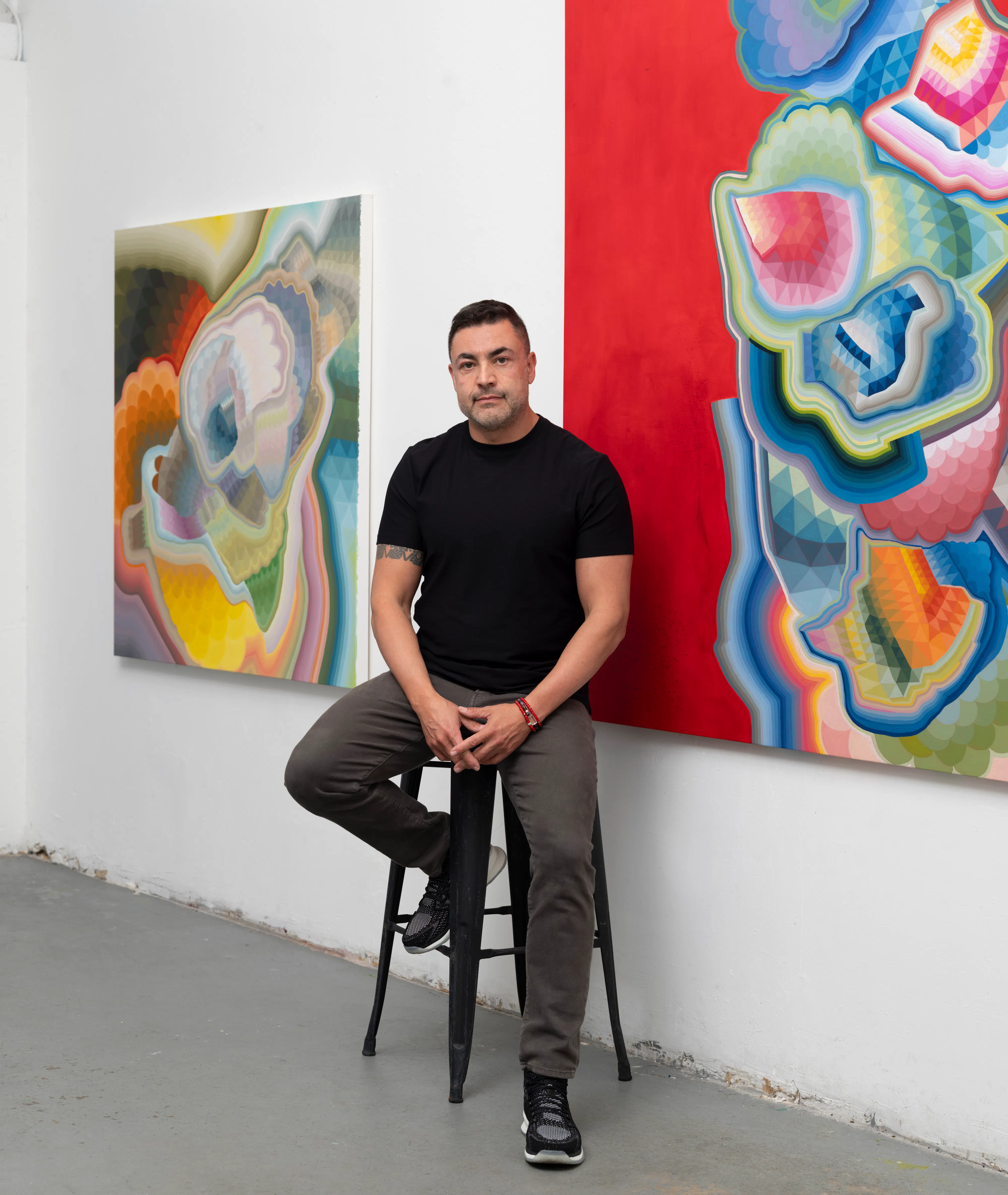 Moncho in his studio, seated on a stool between large-scale abstract paintings, with colorful geometric compositions visible on surrounding walls, emphasizing the artist within his working environment.