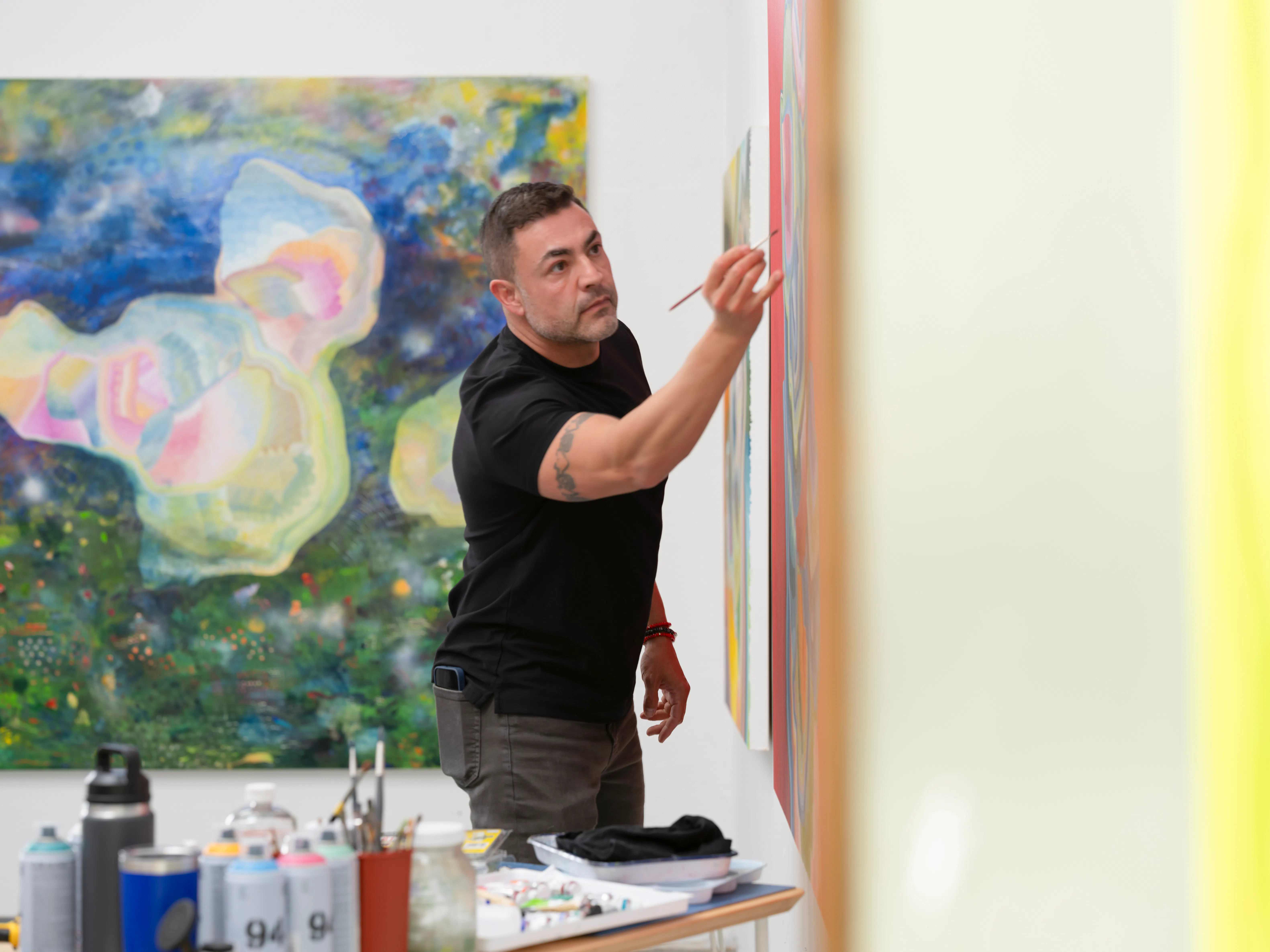 Artist Moncho painting in his studio, applying detail to a canvas with a brush, with tools and materials visible in the foreground and large-scale abstract works in the background.