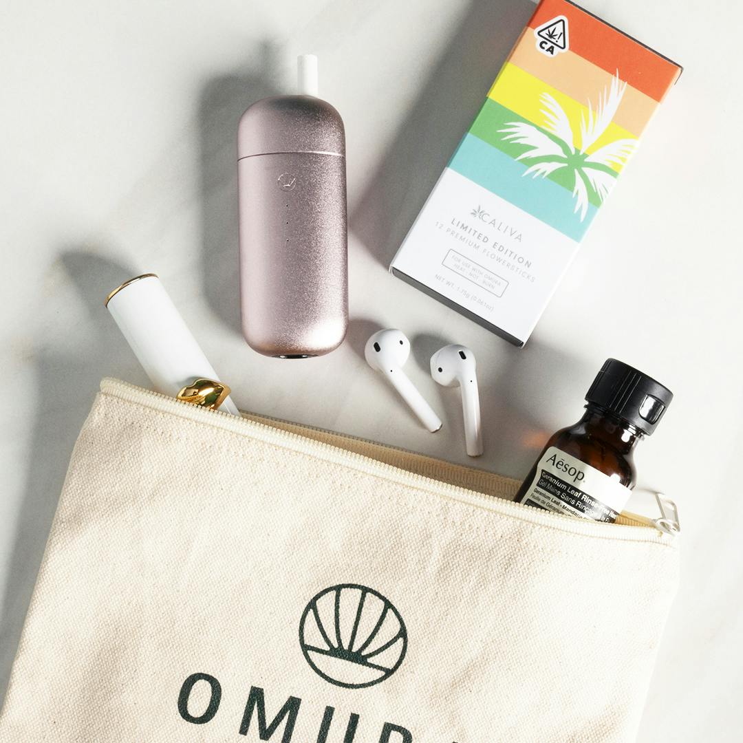 small white bag laying open with caliva flowersticks, omura device, and airpods displayed on flat surface 