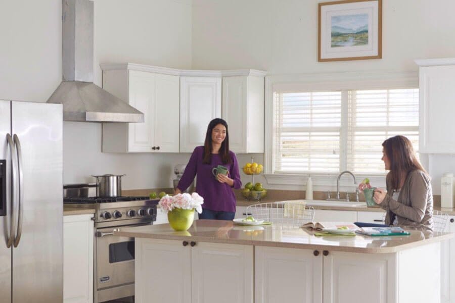 Two women in a brightly lit kitchen with white faux wood blinds in the windows.