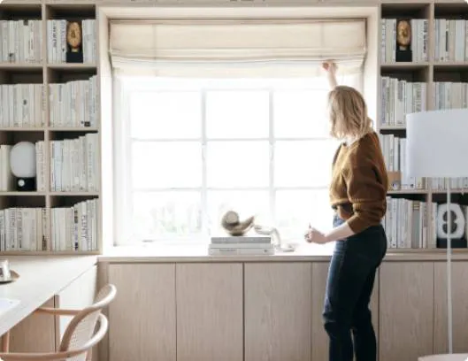 A woman standing by a window, reaching for the window shade and is about to pull it down.