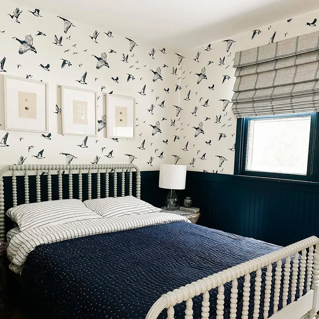 A cream and navy bedroom. The walls have wallpaper with birds on it. The window has gray roman shades with navy details.