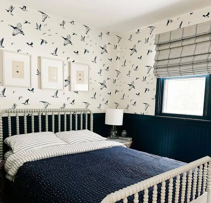 A cream and navy bedroom. The walls have wallpaper with birds on it. The window has gray roman shades with navy details.