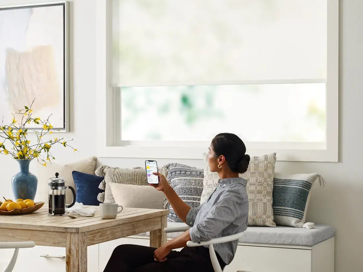 A woman sits at a breakfast nook using an remote controlled app to control white roller shades.