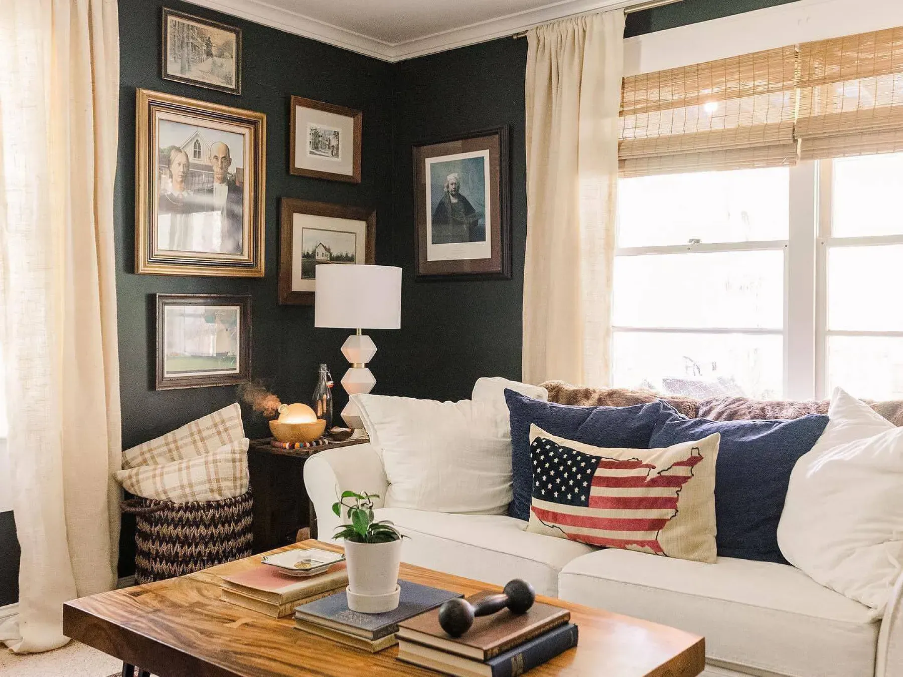 Living room space with dark green walls and a white couch. The windows are covered with sheer curtains layered over light woven wood shades. The wall features a collage of photos and the couch has a pillow with the U.S. on it.