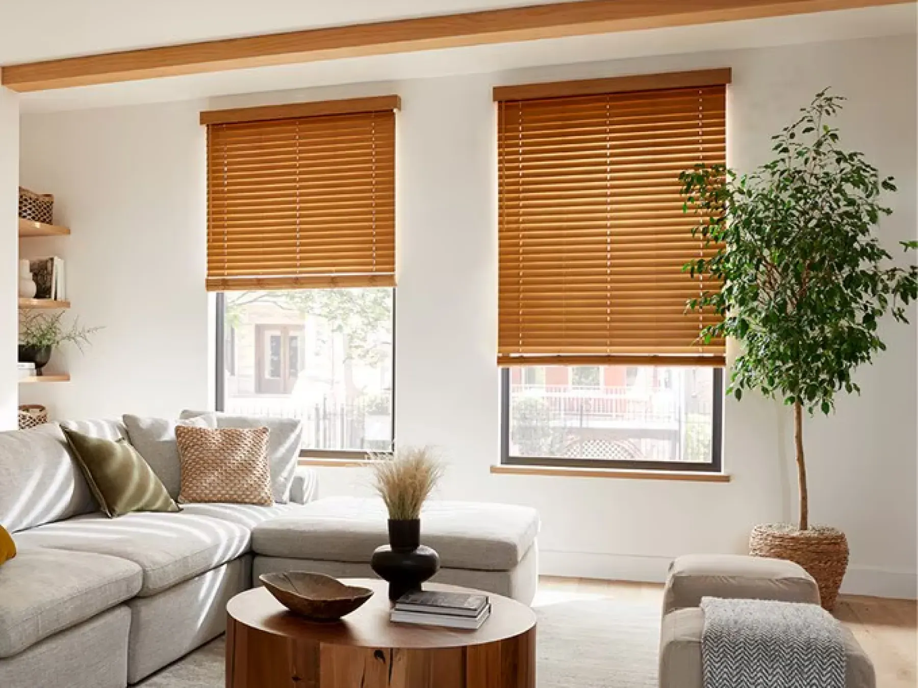 Living room space with a gray couch  and warm-toned wood accents. The windows are covered with raise, brown faux wood blinds. To the right of the windows is an indoor plant. A circular coffee table sits in front of the couch.