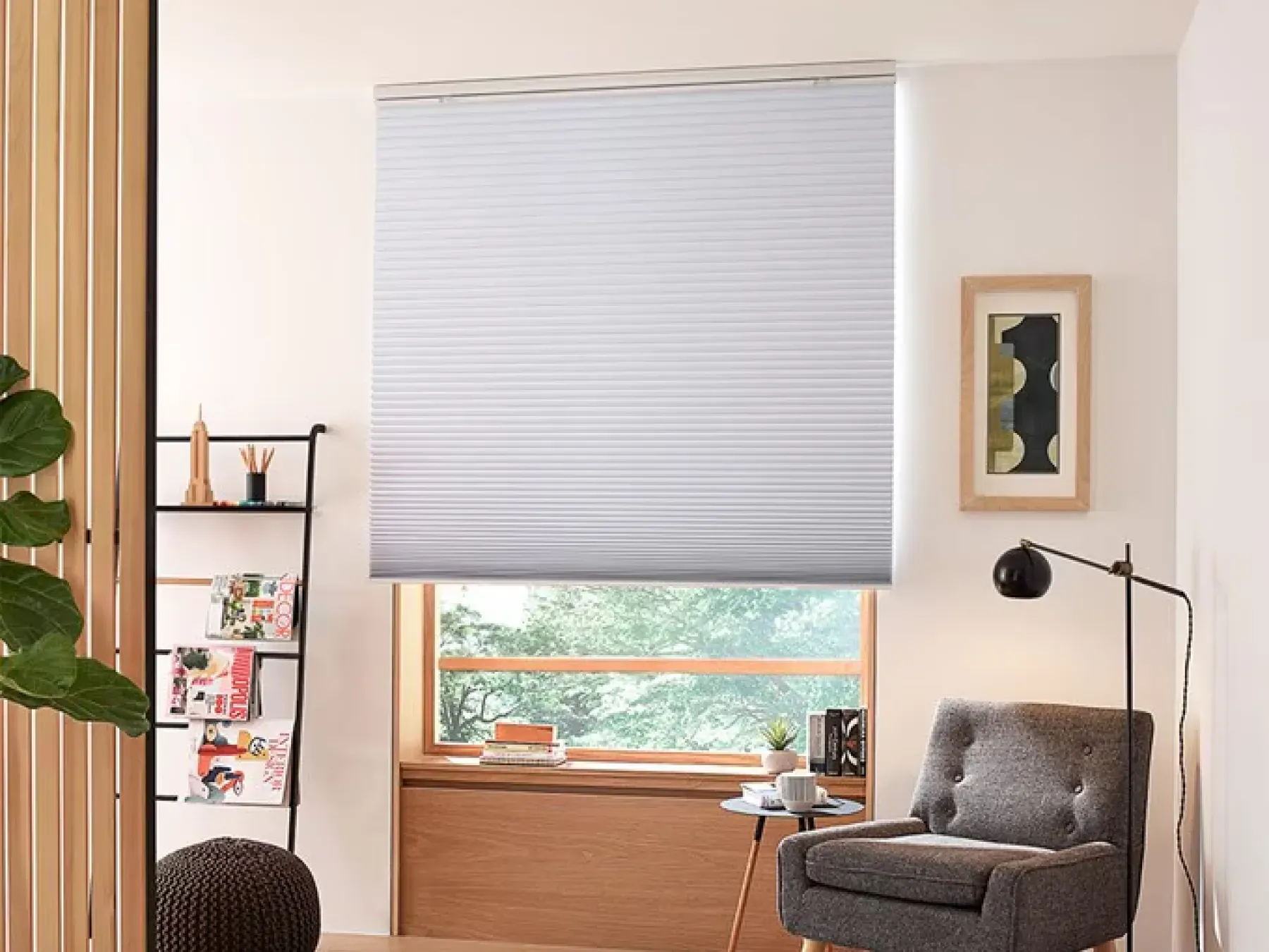 A window with light wood trim and white cellular shades. To the right of the window is a dark gray reading chair and industrial lamp. To the left of the window is a ladder bookshelf with various decorations.