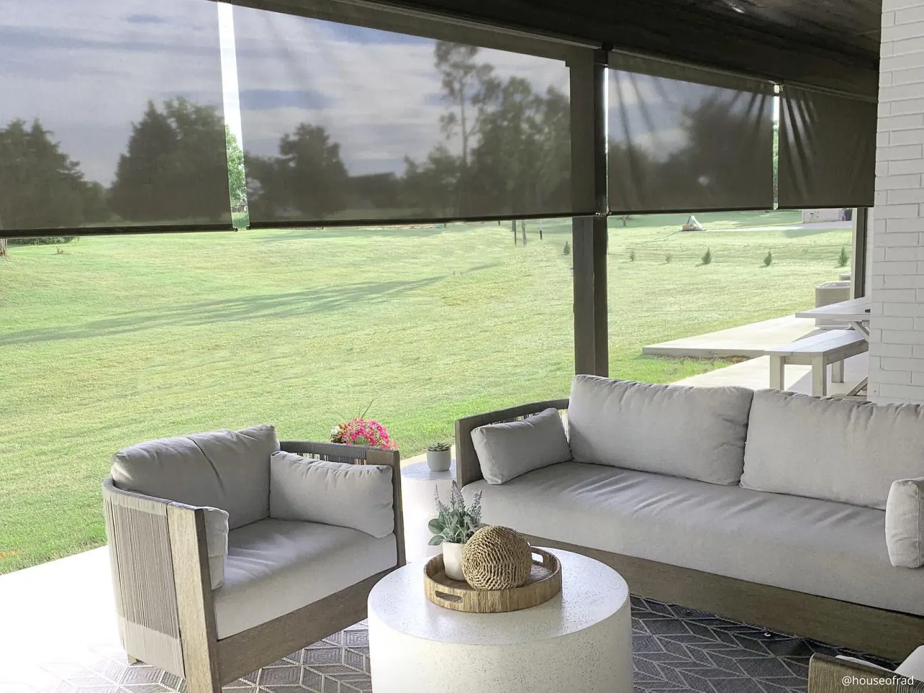 Outdoor sitting space with neutral couches surrounding a round coffee table. Dark colored outdoor shades hang from the beams. In the background is an expanse of grass.