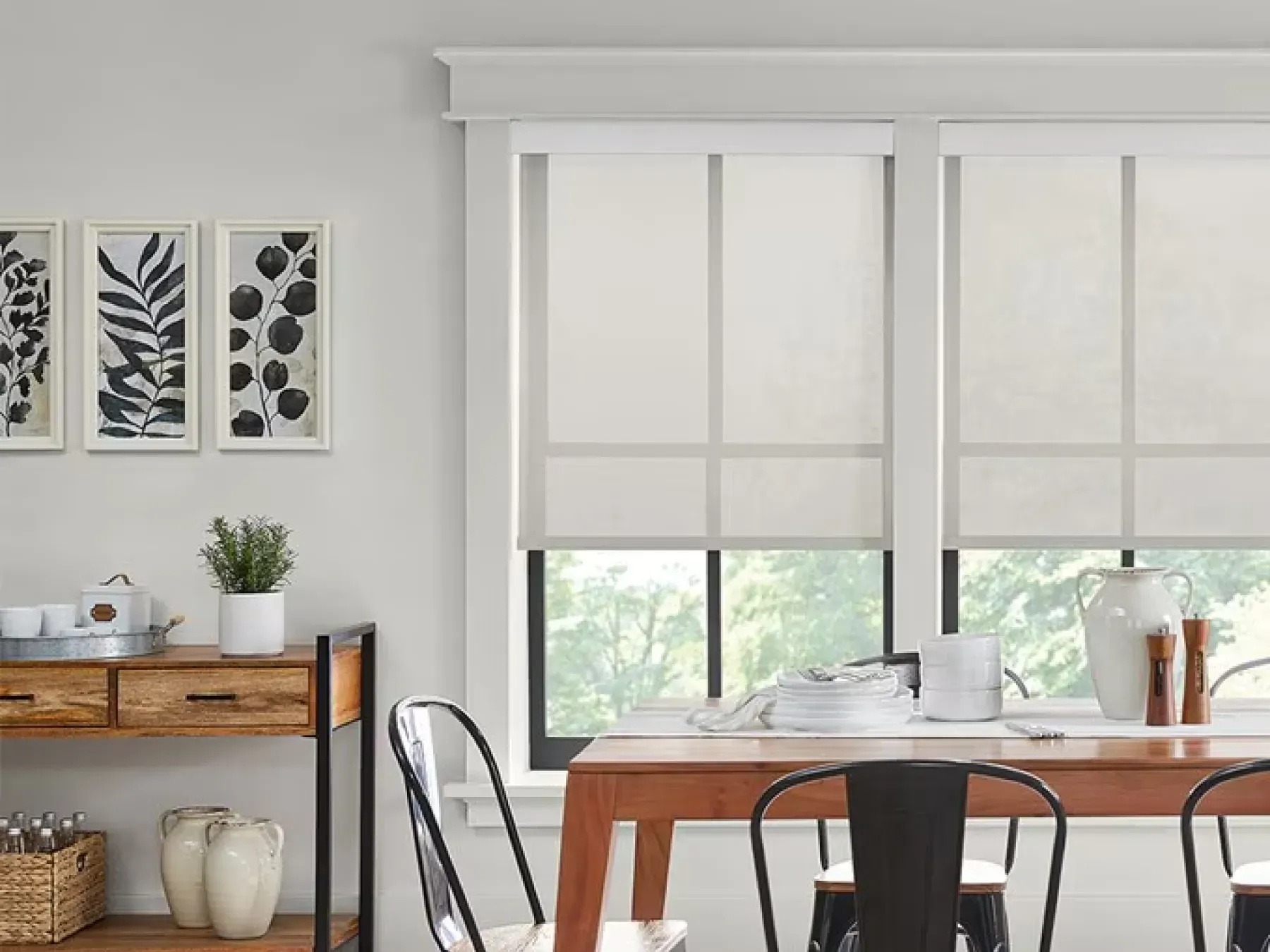 Neutral-colored dining room with white solar shades on the window. The wall to the left of the window features art prints of leaves. Below the prints is small counter with decorations. The table is a natural brown with metal chairs around it.