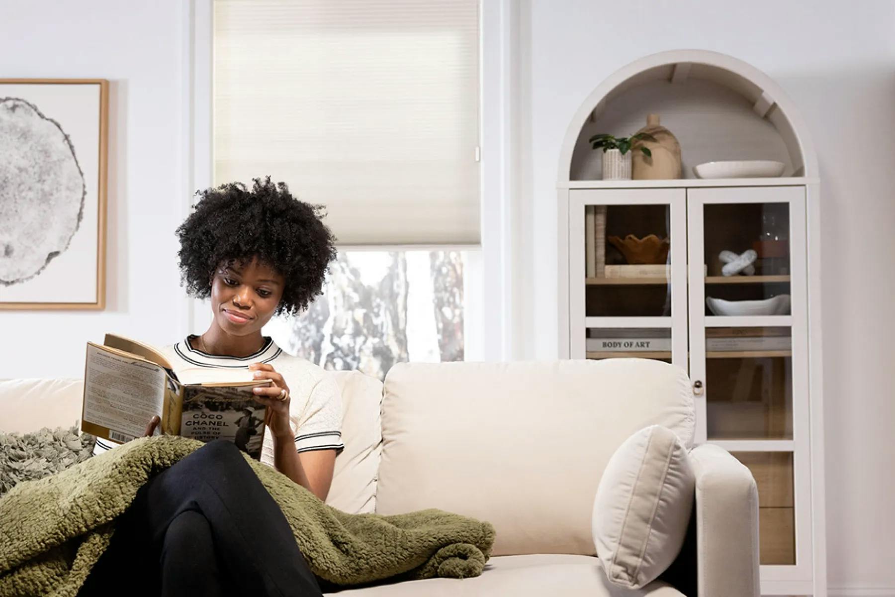 A woman sitting on her couch, reading a book. Behind her is a window covered with white cellular shades. To the right of the window is a bookshelf.