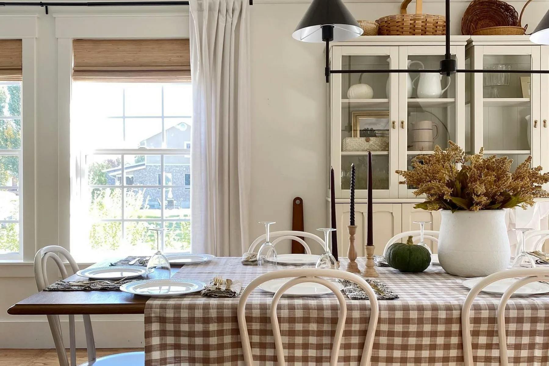 Dining room with earth-tone furniture, a white china cabinet, windows with woven wood shades and white curtains. The table is set for a fall dinner.