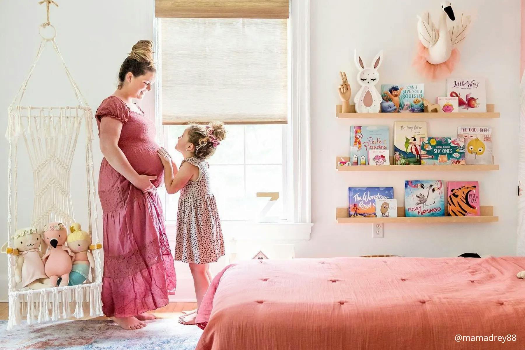 A pregnant woman standing with her daughter in front of a window. The window is covered with woven wood shades.