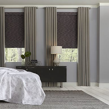 Bedroom with elegant floor-length gray drapes layered over textured blackout roller shades, styled beside a modern black console table and lamp