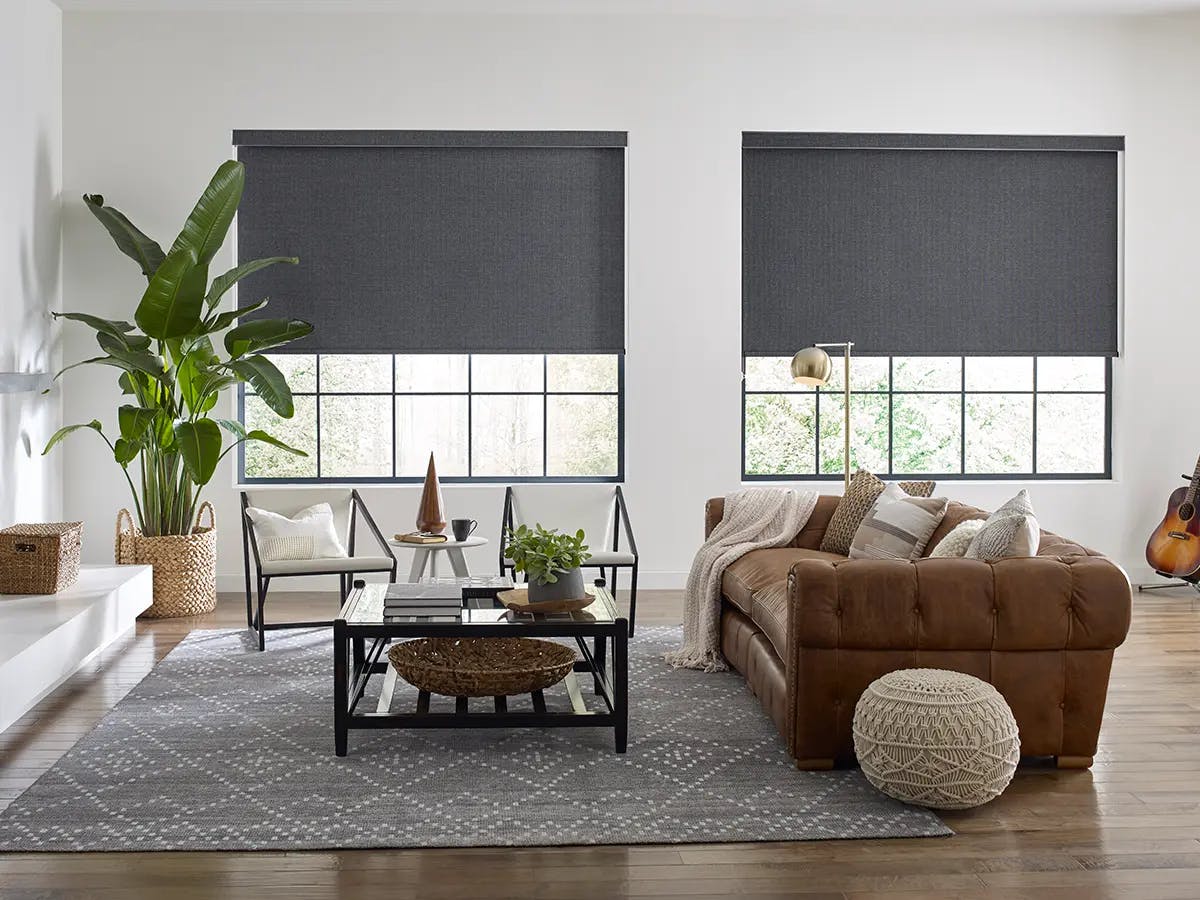 A cozy living room with a brown leather sofa, large windows with dark roller shades and a gray patterned rug.