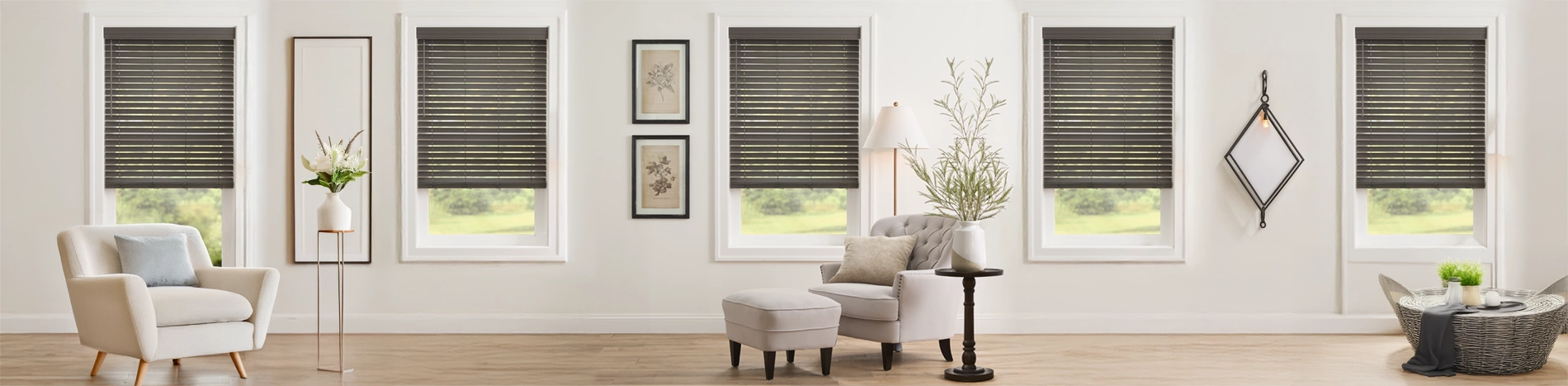 Warm and inviting living room scene featuring multiple windows dressed in stylish Grey Faux Wood Blinds, providing excellent light control and a modern aesthetic. The space is beautifully furnished with a cream armchair, a tufted side chair with a matching ottoman, and natural decor elements like potted plants and a woven coffee table.
