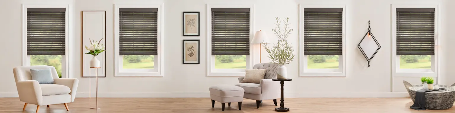 Warm and inviting living room scene featuring multiple windows dressed in stylish Grey Faux Wood Blinds, providing excellent light control and a modern aesthetic. The space is beautifully furnished with a cream armchair, a tufted side chair with a matching ottoman, and natural decor elements like potted plants and a woven coffee table.