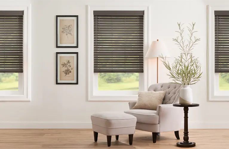 Warm and inviting living room scene featuring multiple windows dressed in stylish Grey Faux Wood Blinds, providing excellent light control and a modern aesthetic. The space is beautifully furnished with a cream armchair, a tufted side chair with a matching ottoman, and natural decor elements like potted plants and a woven coffee table.