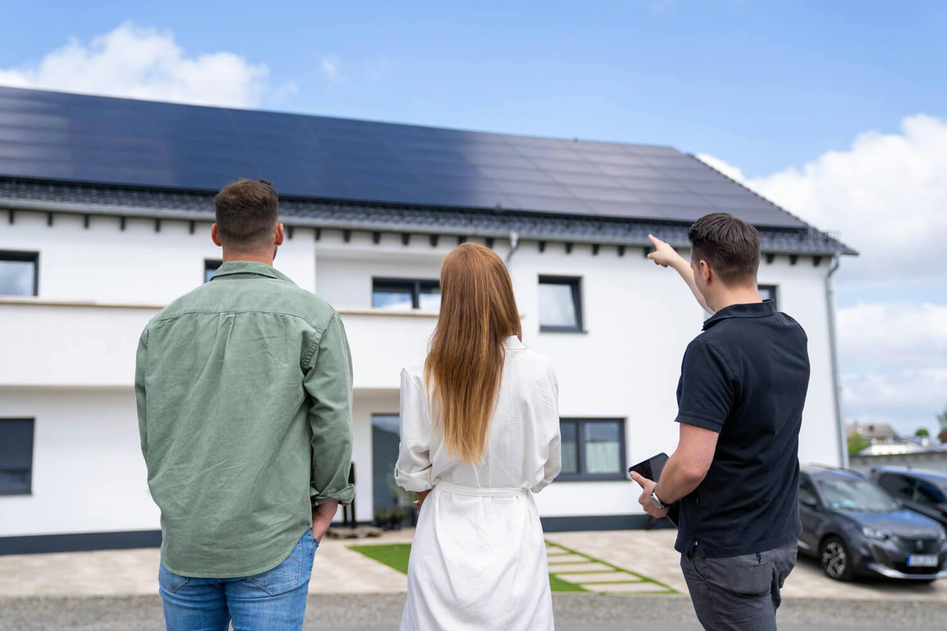 Drei Personen stehen vor einem modernen Haus mit Photovoltaikanlage auf dem Dach, während ein Berater auf die Solarmodule zeigt.