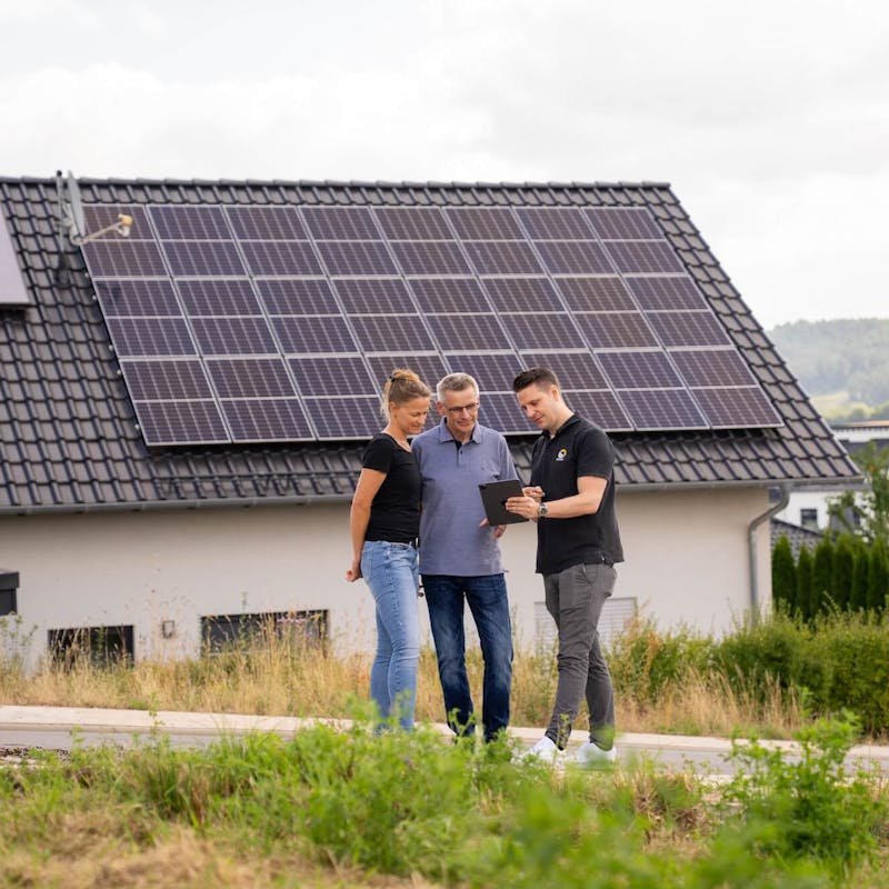 Paar wird vor Haus mit Solaranlage auf dem Dach von einem Gelb Solar Experten zu Stromspeicher beraten Paar wird vor Haus mit Solaranlage auf dem Dach von einem Gelb Solar Experten zu Stromspeicher beraten