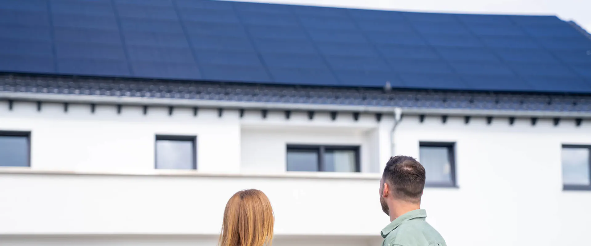 Paar steht vor seinem Einfamilienhaus mit Photovoltaikanlage auf dem Dach und betrachtet die neue Solaranlage Paar steht vor seinem Einfamilienhaus mit Photovoltaikanlage auf dem Dach und betrachtet die neue Solaranlage