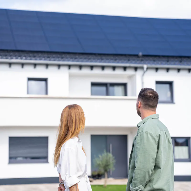 Paar steht vor seinem Einfamilienhaus mit Photovoltaikanlage auf dem Dach und betrachtet die neue Solaranlage Paar steht vor seinem Einfamilienhaus mit Photovoltaikanlage auf dem Dach und betrachtet die neue Solaranlage