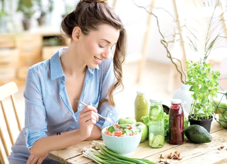 Genç kadın yemek masasında salata yiyor ve gülümsüyor. Yemek masası üzerinde avokado, soğan, limonlu su, elma ve fındık bulunuyor.