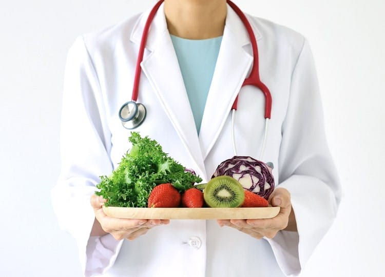 Yakın plan çekimde, beyaz doktor önlüğü giyen kadın elinde tahta bir tabak tutuyor. Tabağın üzerinde; kivi, çilek, maydanoz ve kırmızı lahana bulunuyor.