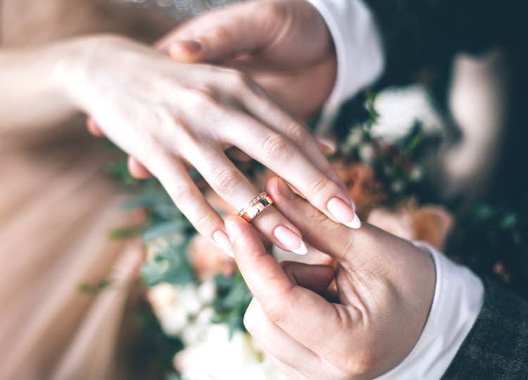 Yakın plan çekimde; adam kadının yüzük parmağına altın rengi bir alyans takıyor. Kadının diğer elinde bir çiçek buketi bulunuyor.