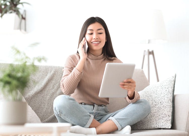Genç kadın oturma odasındaki açık renkli koltuğunda otururken; sol eli ile tabletine bakarken, sağ ile ile telefon görüşmesi yapıyor ve gülümsüyor.