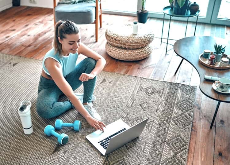 Genç kadın salondaki zeminde bulunan kahverengi halı üzerinde bağdaş kurarak otururken, önünde duran laptopa bakarak gülümsüyor ve sağ eli ile laptopa dokunuyor. Kadının sağında su şişesi, iki adet küçük dambıl bulunuyor. 