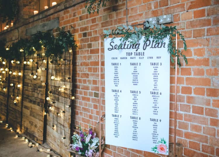 Düğün salonunun girişinde, dekoratif olarak ışıklandırılmış paneller, çiçek buketleri ve masa numaralarının altına isimlerin yazılmış olduğu büyük bir oturma planı tablosu bulunuyor.