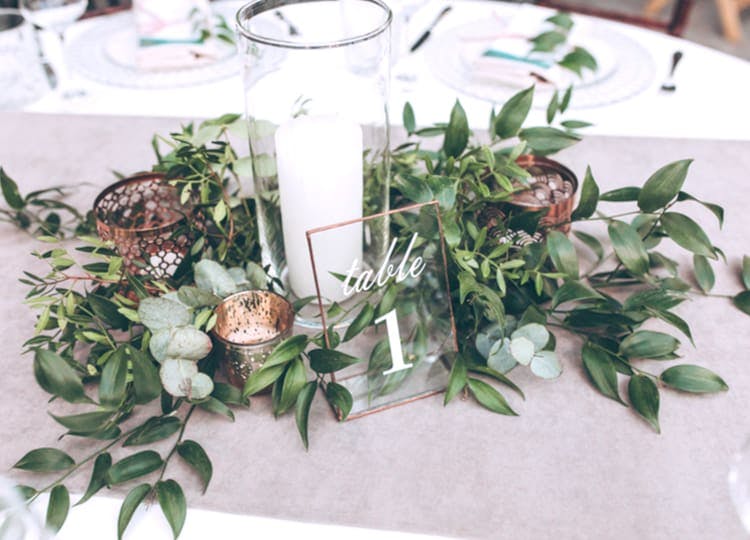 Yemek masası üzerinde; tabaklar, bardaklar, çatallar, dekoratif masa çiçeği ve çiçeğin ortasında bir mum duruyor. Mumun önünde ise masa numarası bulunuyor.