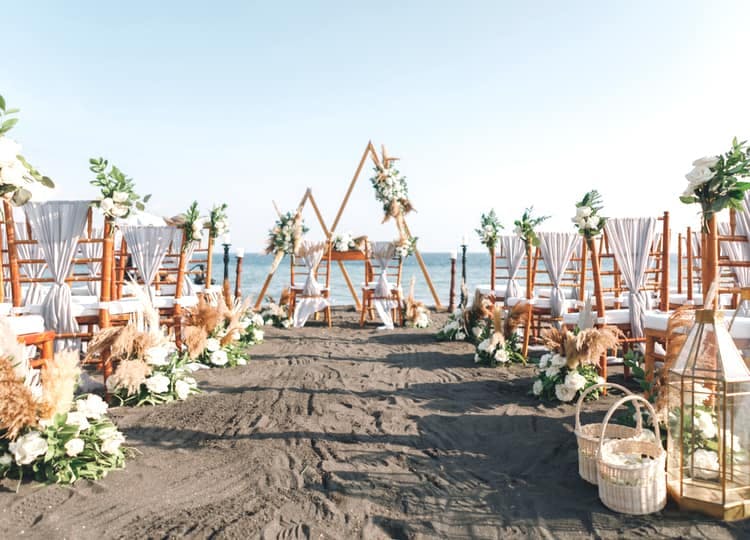 Kumsalda dekor olarak üçgen tahtalar, çiçek buketleri ve beyaz tüller ve çiçekler ile süslenmiş sandalyeler bulunuyor.
