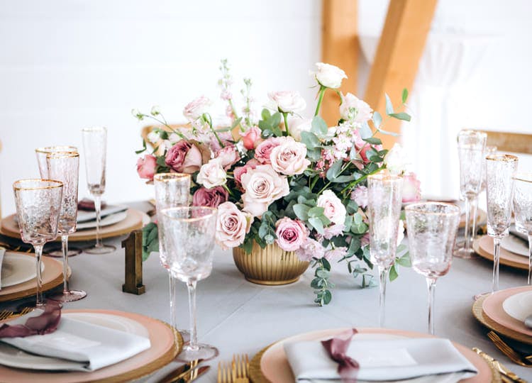 Düğün salonunda yemek masası üzerinde tabaklar; bardaklar, çatallar, masa numarası ve çiçek buketi bulunuyor.