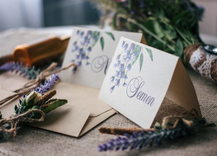 Koyu renk arka plan üzerinde; ikiye katlanmış düğün davetiyeleri, davetiye zarfı, bir buket çiçek ve masaya serpiştirilmiş lavanta dalları bulunuyor.