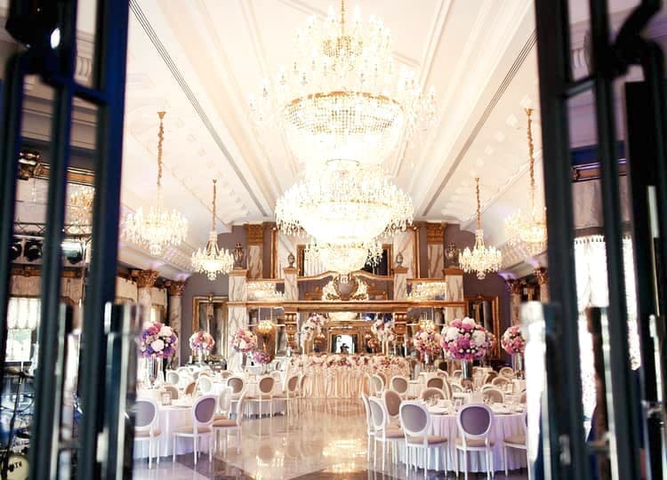 Düğün salonu içerisinde yuvarlak yemek masaları, sandalyeler, masaların üzerinde dekoratif çiçekler, büyük ve gösterişli avizeler bulunuyor.