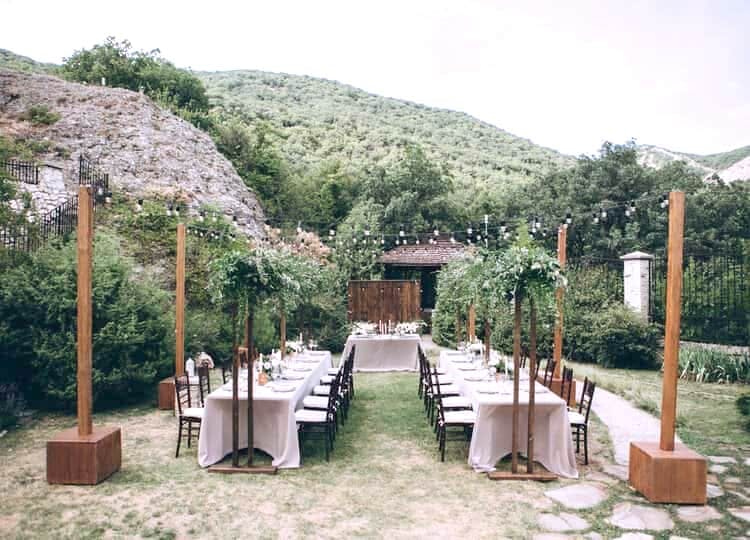 Rustik temalı olarak yapılan düğünde, bahçe içerisinde iki yemek masası ve dekoratif çiçekler bulunuyor. Masaların üzerinde tabaklar ve çiçek demetleri duruyor. 