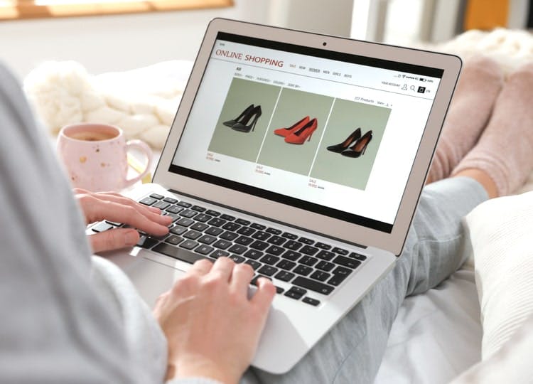 Yakın plan çekimde kadın yatağında ayaklarının uzatarak oturuyor ve dizlerinin üstünde bulunan laptop ekranında topuklu ayakkabı modelleri bulunuyor.