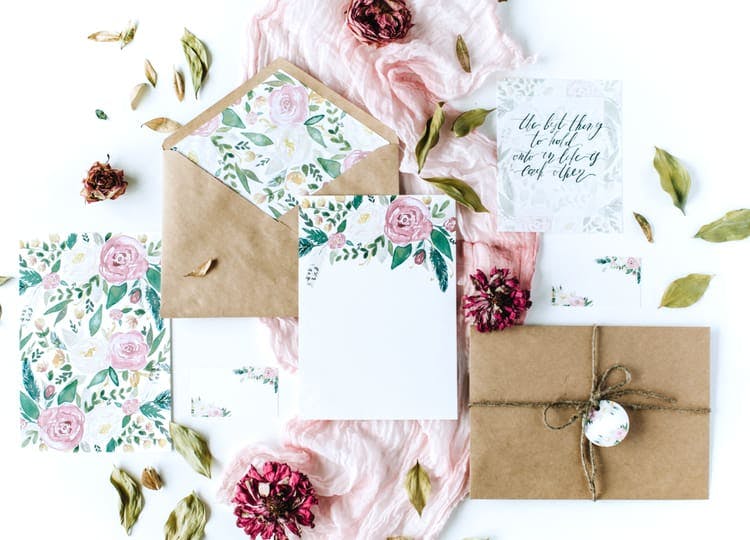 Açık renk arka plan üzerinde; davetiye zarfı, çiçekli davetiyeler, pembe bir tül ve kurumuş çiçek parçaları bulunuyor.
