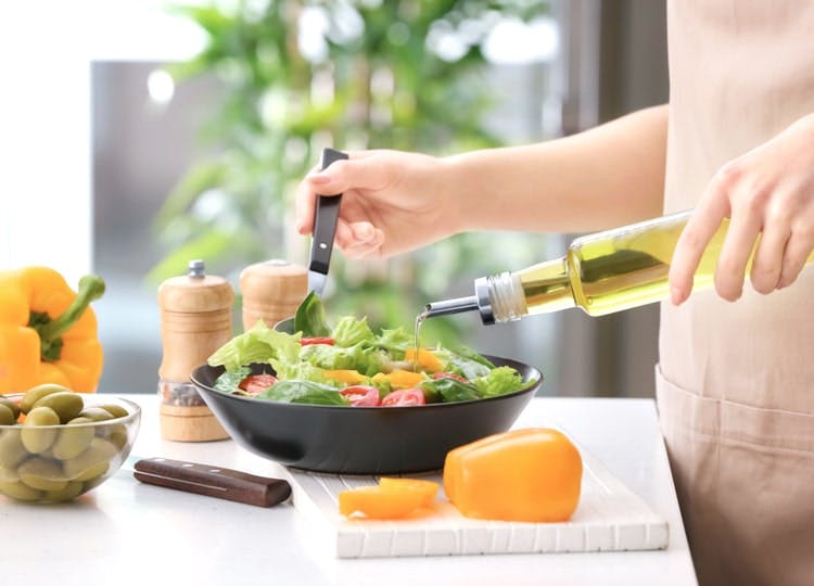 Yakın plan çekimde; kadın mutfaktaki tezgah üzerinde hazırlamış olduğu salata içerisine zeytin yağı döküyor. Tezgahta kesme tahtası, turuncu kapya biberi, yeşil zeytinler, bıçak ve baharatlıklar bulunuyor.