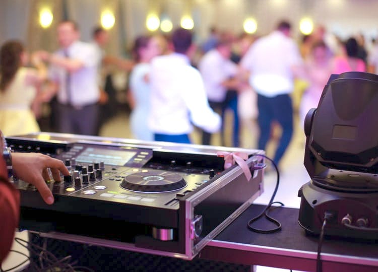 Düğün salonunda yakın plan çekimde DJ ve DJ kabini bulunuyor. Kabinin ön kısmında ise konuklar sahnede dans ediyor.