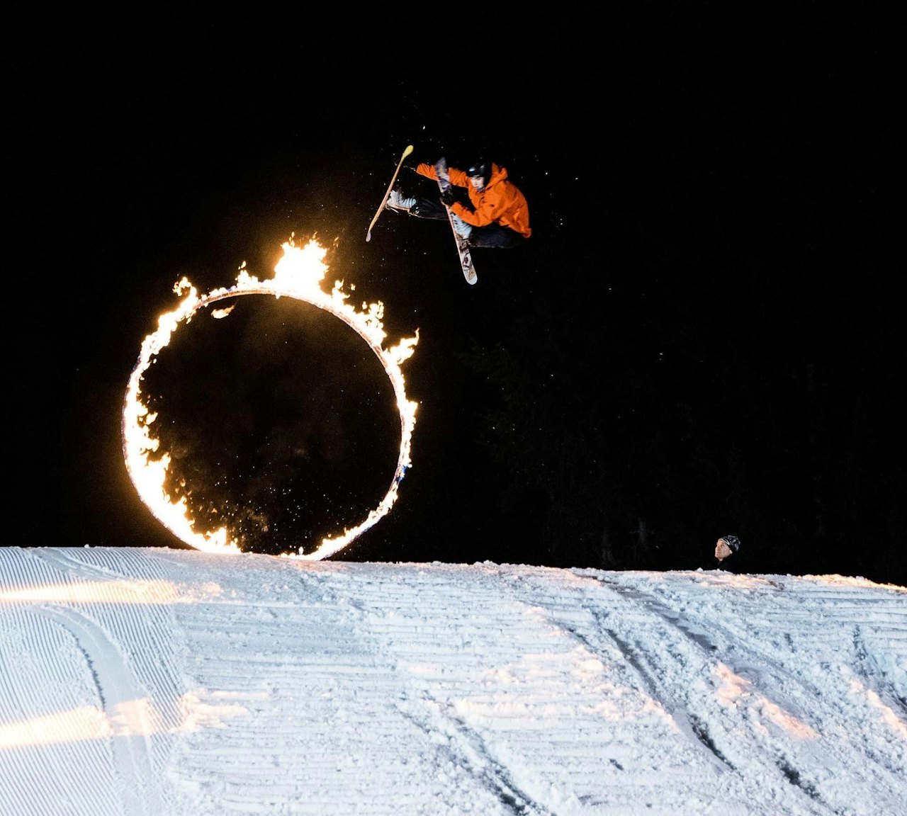 Skier of undiscernible gender in an orange ski jacket skiing through a hoop on fire at night at Whistler Blackcomb in British Columbia, Canada.