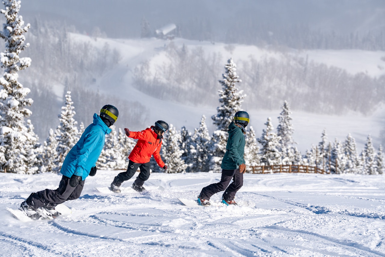 A snowboard instructor in red ski jacket imparting a snowboarding lesson to two young snowboarders of undiscernible gender in colorful snowboarding attire at Snowmass in Colorado, USA.