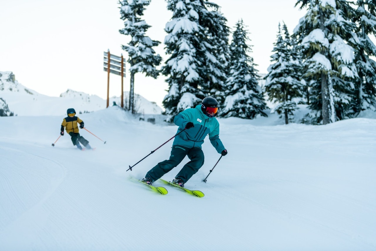 Male ski guide in blue ski jacket giving a lesson to a child of undiscernible gender in yellow ski jacket at Whistler Blackcomb in British Columbia, Canada.