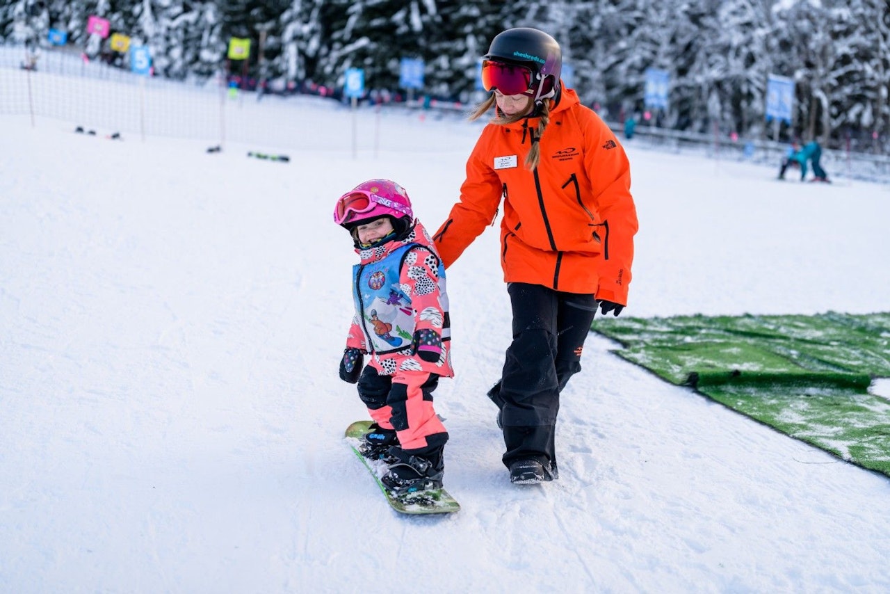 Snowboard guide imparting a snowboarding lesson to a young child of undiscernible gender in pink and blue ski attire at Whistler Blackcomb in British Columbia, Canada.