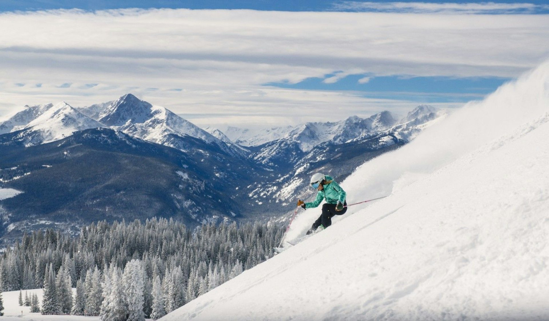 A skier gracefully descends a snowy mountain slope, surrounded by tall pine trees and a clear blue sky.